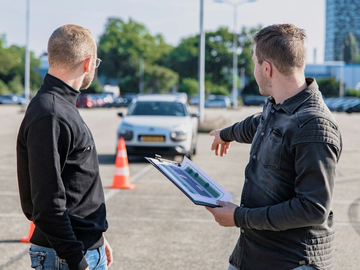 En kørelærer gennemgår øvelsen med en elev på pladsen, mens en bil holder klar ved keglerne.